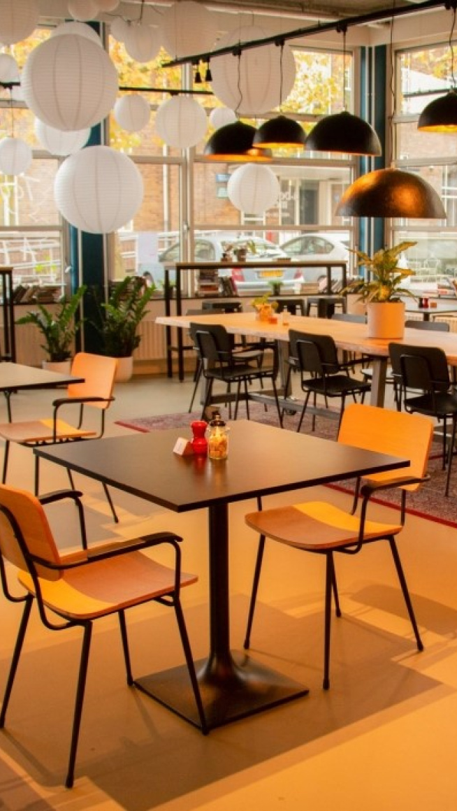 Ontmoetingsruimte A Beautiful Mess met gele vloer, een vloerkleed, tafels en stoelen, ramen rondom en aan het plafond witte lampions en een aantal zwarte lampen