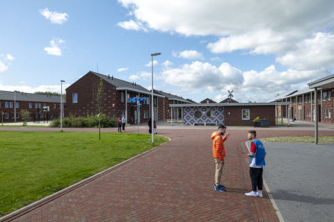2 pratende mensen staan op een terrein met daarachter gebouwen van twee verdiepingen met puntdak