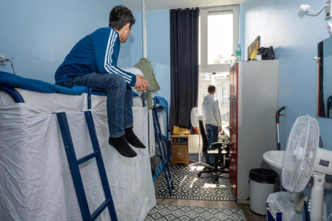 twee jongeren waarvan 1 op bed zit en naar buiten kijkt en de andere staat voor het raam