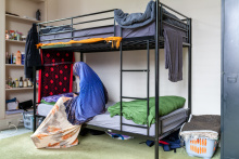 Een amv meisje zit in haar slaapkamer op het onderste bed van een stapelbed en kijkt op haar telefoon. Je ziet haar vanaf de linkerzijkant. Ze draagt een blauwe hijab en een lange jurk. De open kast links achter het bed staat vol met spulletjes waaronder shampoo. Er staat een volle wasmand rechts naast het bed. Aan het bed hangen handdoeken te drogen.