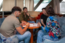 Drie jonge mannelijke statushouders zijn in gesprek met een vrouwelijke woonbegeleider in de salon van het schip waar zij verblijven. De woonbegeleider vertelt iets waar de mannen om moeten lachen. Ze zitten op blauwe banken rondom een ronde houten tafel. Er liggen papieren op tafel en de linkerman houdt een pen vast.