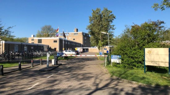 ingang azc met slagboom en COA bord met naam azc daarachter gebouwen, gras, struiken en bomen